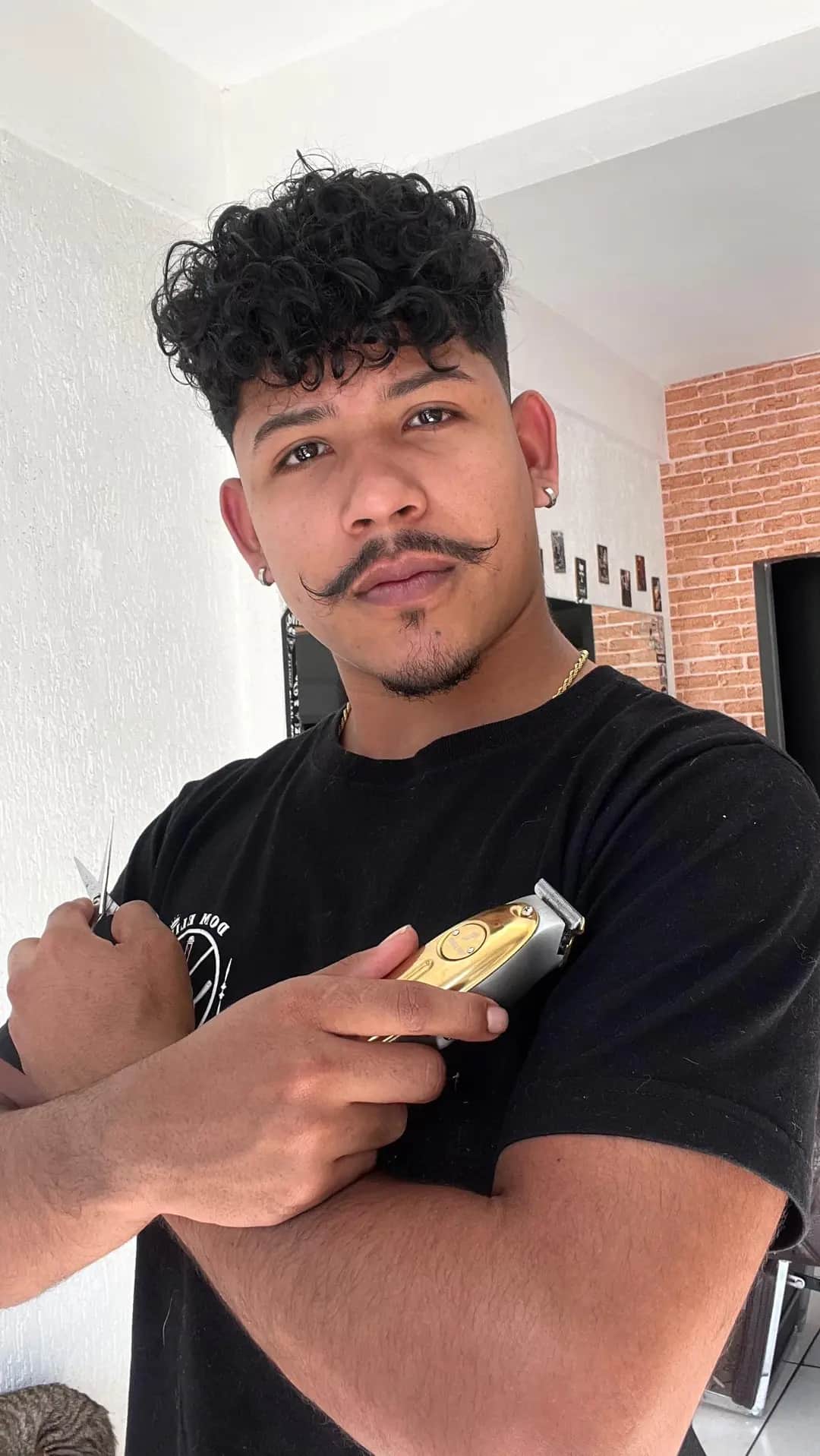 Barbeiro da Dom Elii Barbershop com máquina e tesoura na mão, cabelo cacheado com degradê nas laterais